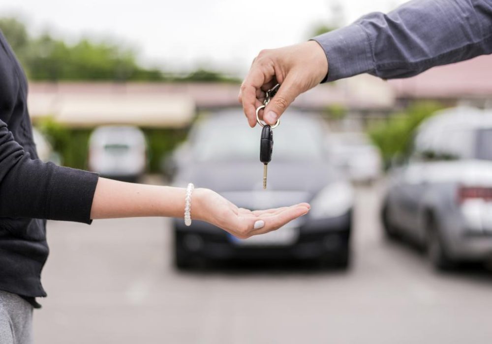transportation and ownership concept - customer and salesman with car key outside. Woman at a car dealership buying an auto, the sales rep giving her the key, macro shot with focus on hands and key